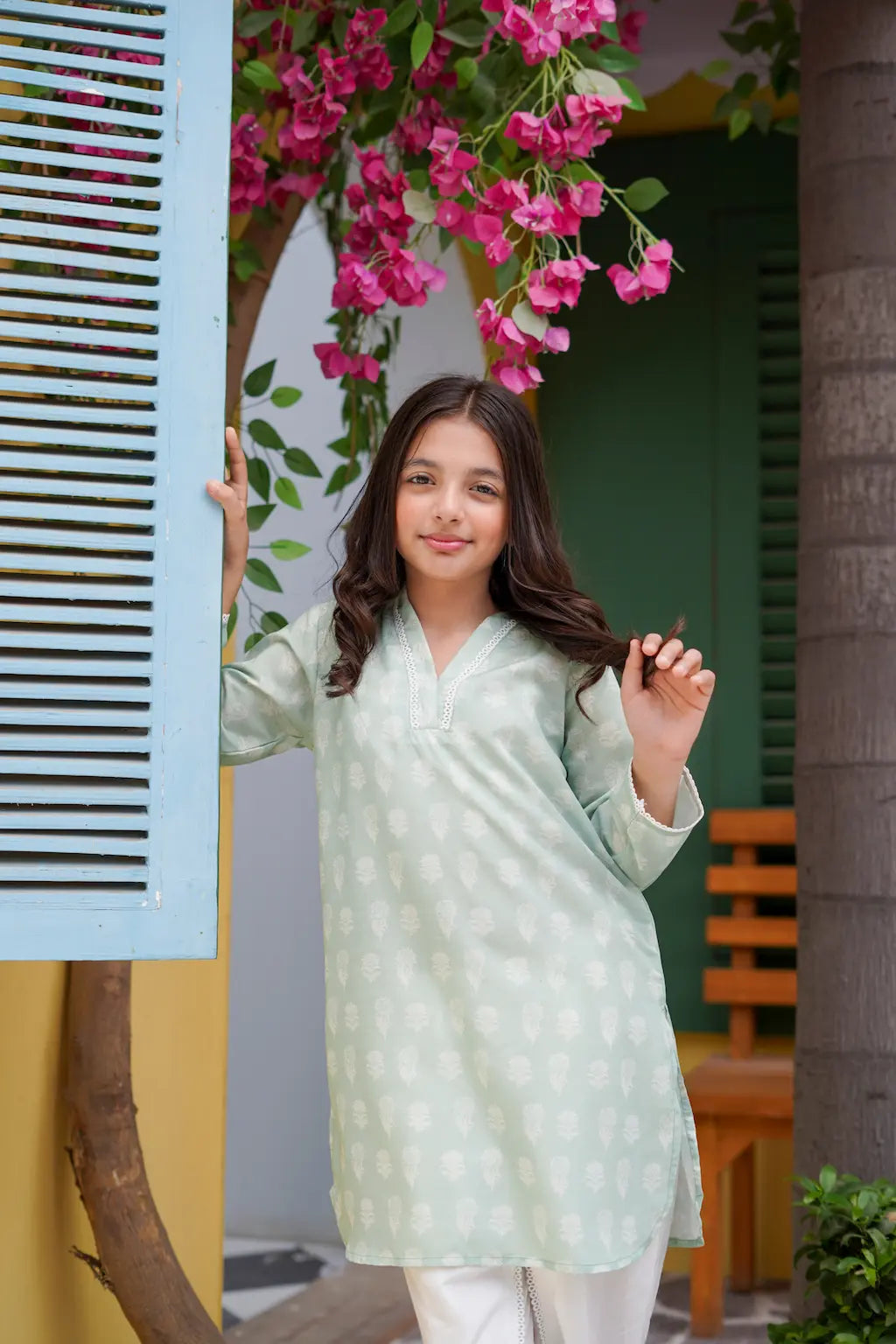 Young girl in a light green traditional outfit standing outdoors with pink flowers above her head.