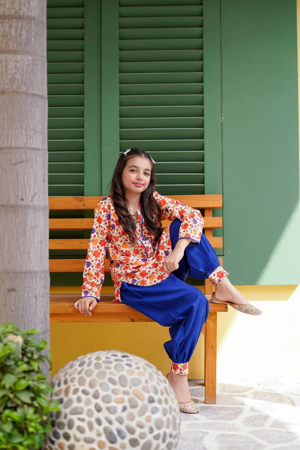 A young girl sitting on a wooden bench in front of green shutters