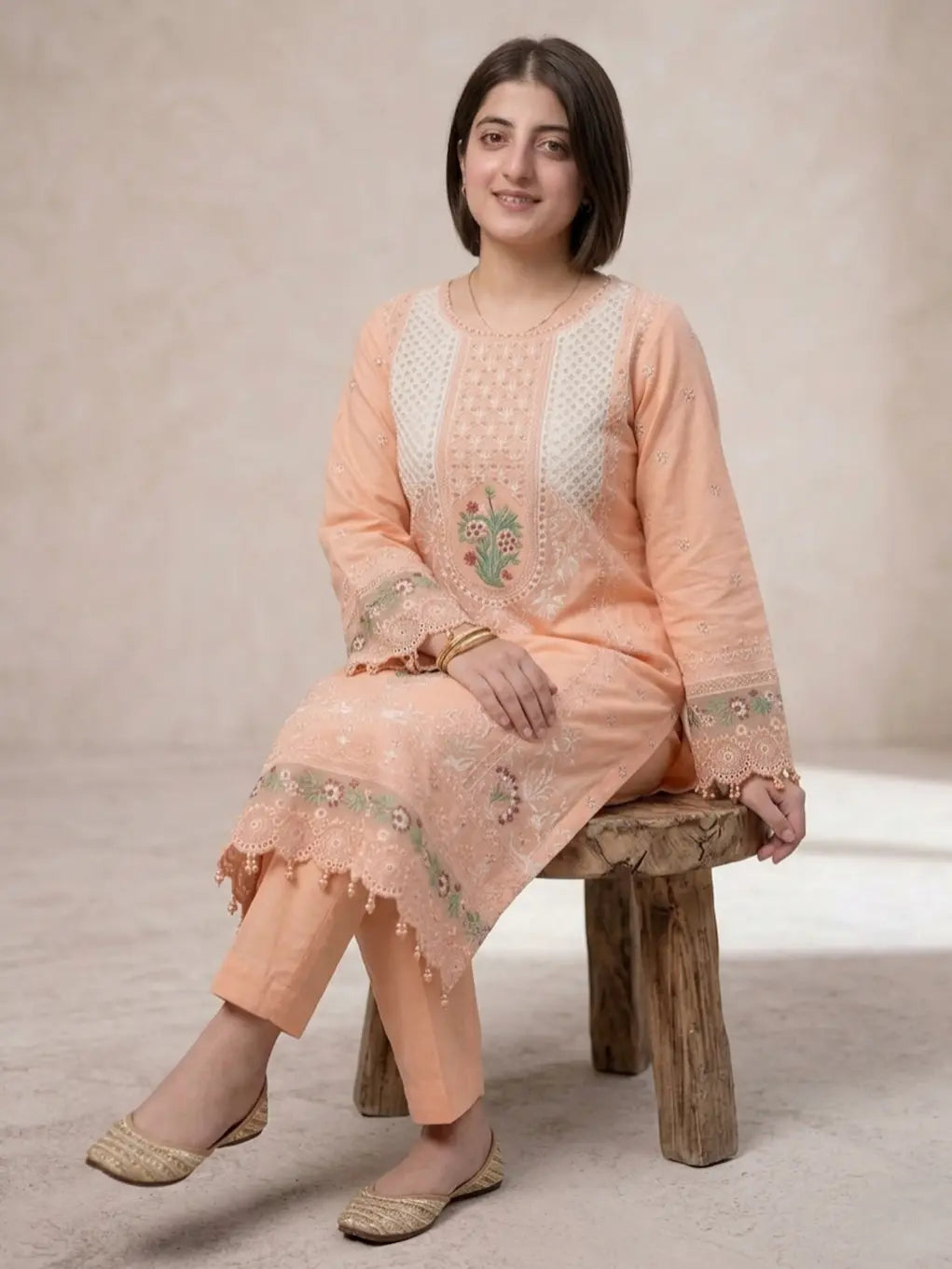 Woman in peach embroidered traditional outfit sitting on a wooden stool against a neutral background