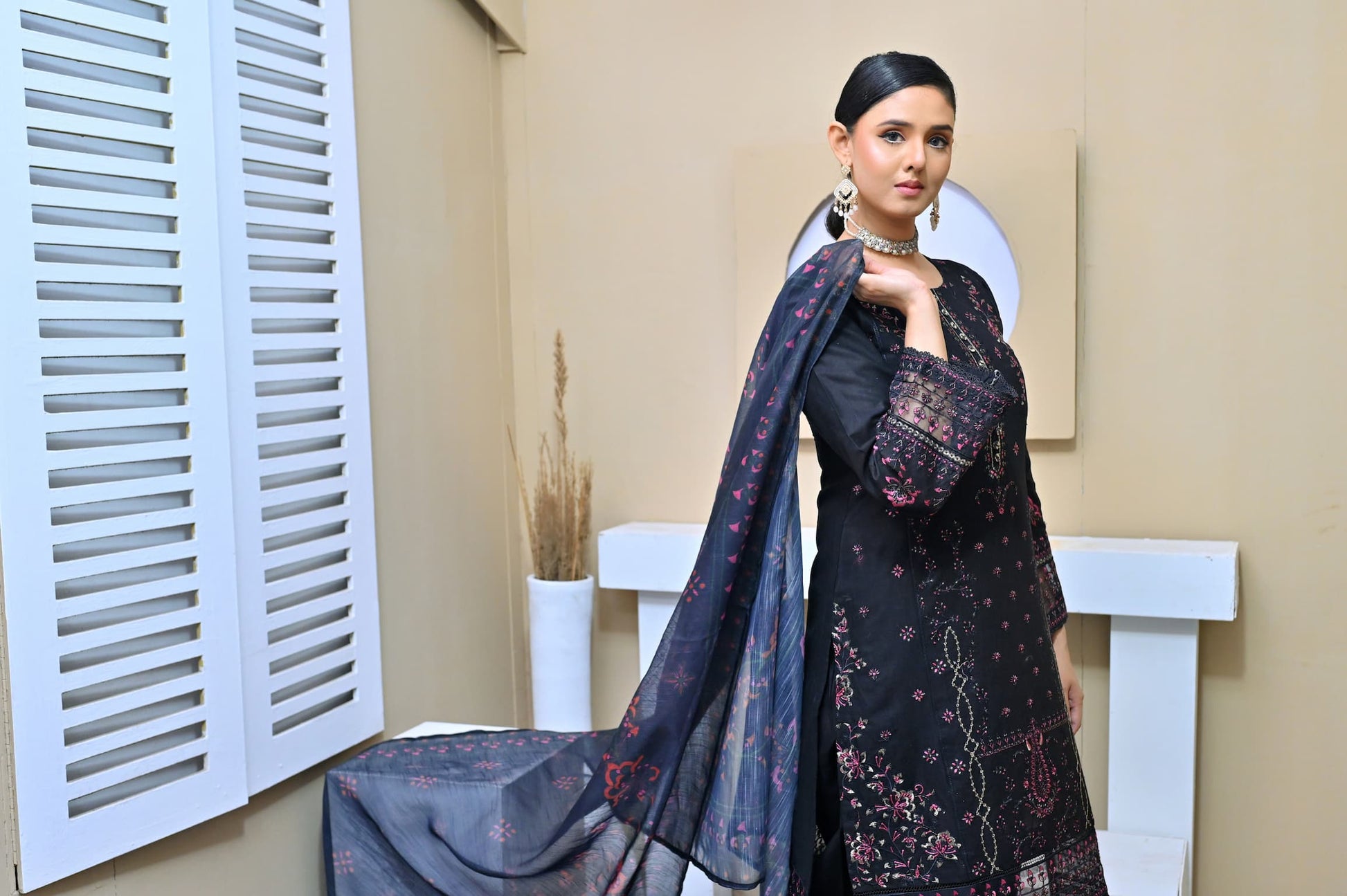 Model in a black three-piece embroidered suit with pink floral details, lace-trimmed sleeves, and a printed dupatta, posing in a minimalist studio setting.