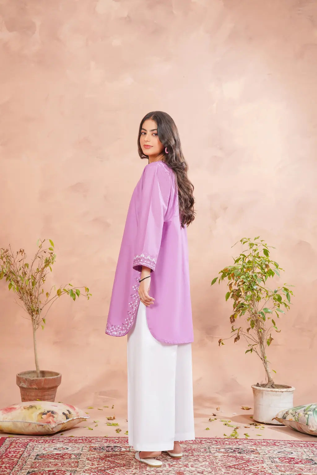 Woman wearing a light purple top and white pants against a beige wall with plants.