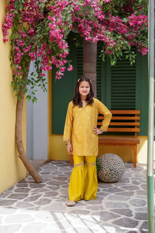 Young girl in a yellow outfit standing in front of a yellow wall with pink flowers and a wooden bench.