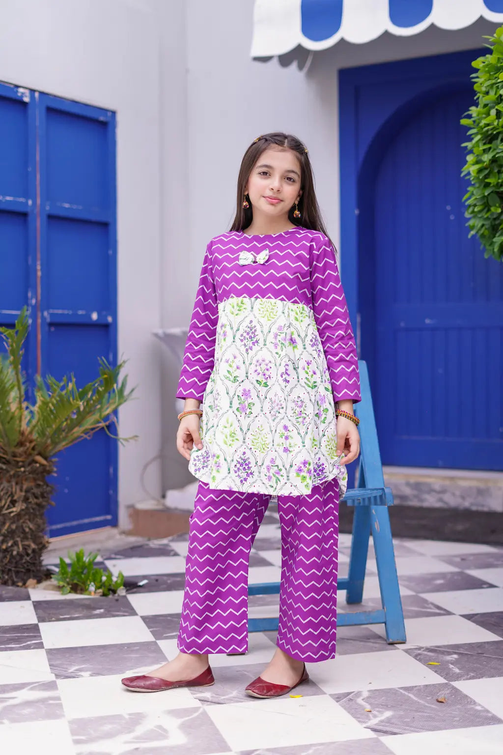 Young girl in a purple and white patterned outfit standing in front of a blue door.