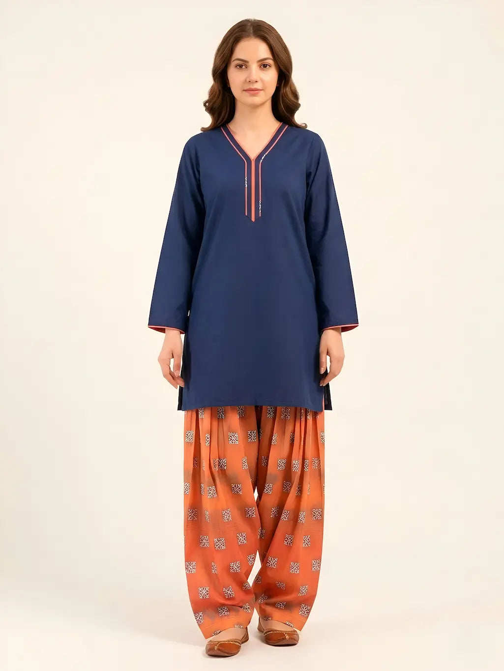 A woman posing in a navy blue short-length lawn tunic with coral-pink piping on the V-neckline and cuffs, paired with a voluminous, digital-printed rust Farshi shalwar and traditional brown flats.