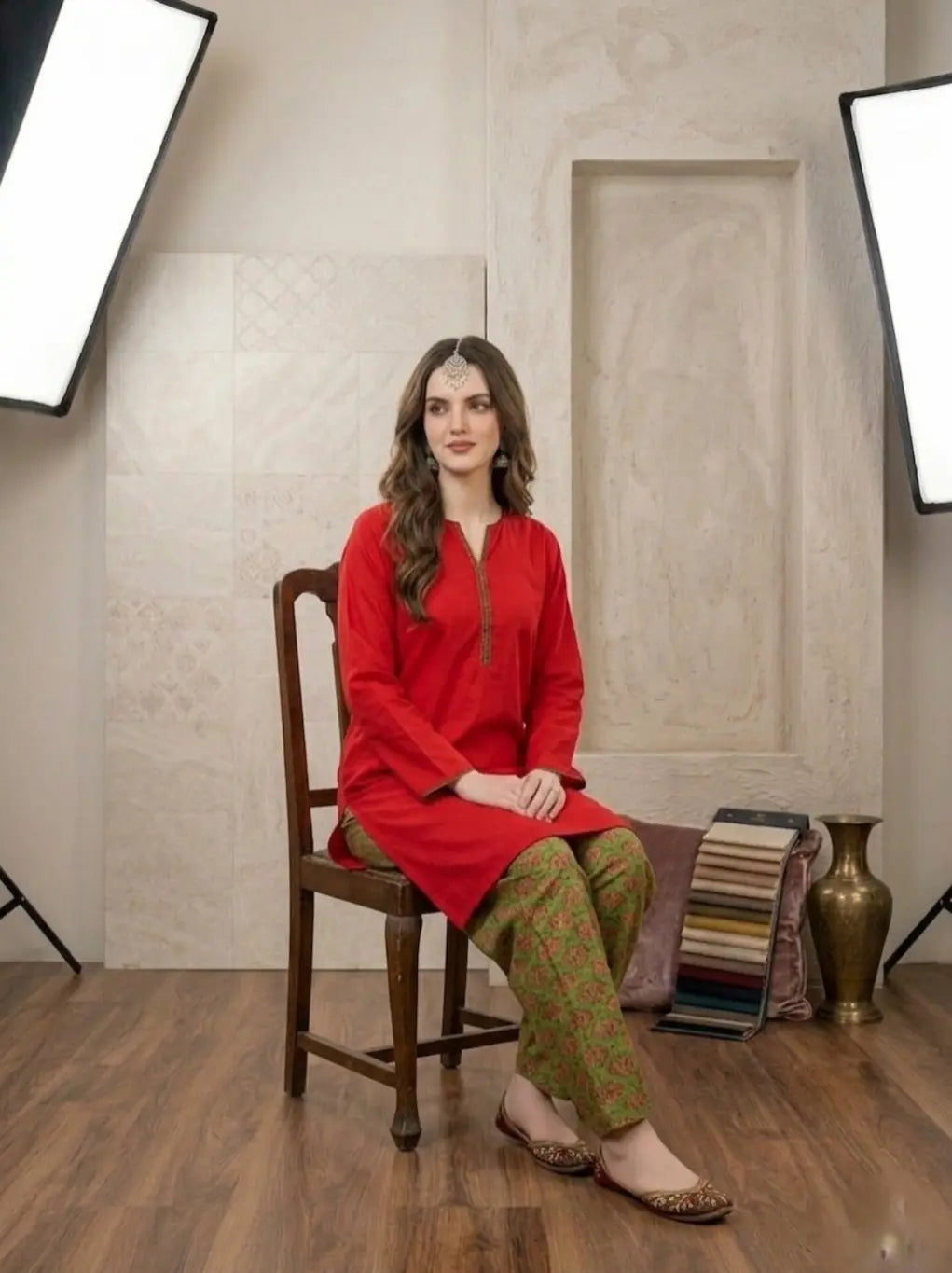 Woman in a red top and green printed shalwar sitting on a wooden chair in a studio setting.