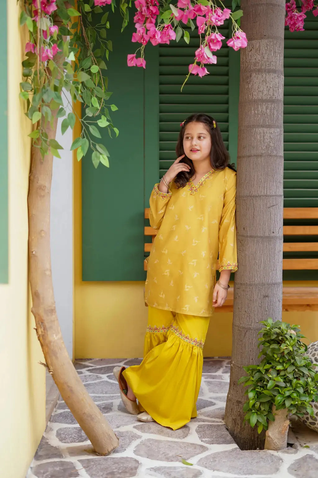 A young girl in a yellow traditional outfit standing under a tree with pink flowers.