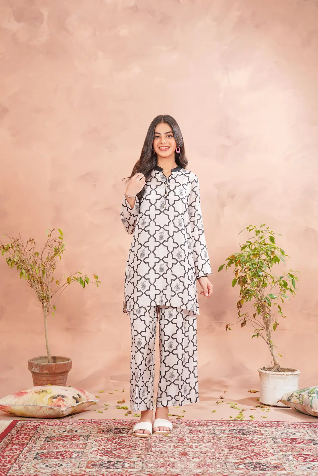 Woman wearing a patterned outfit standing against a pink wall with plants.