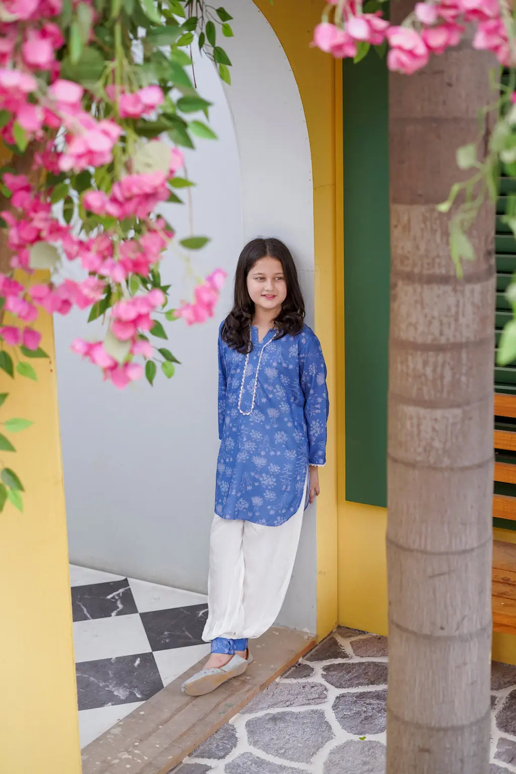 Young girl in a blue top and white pants standing in a decorative outdoor setting with pink flowers and a yellow arch.