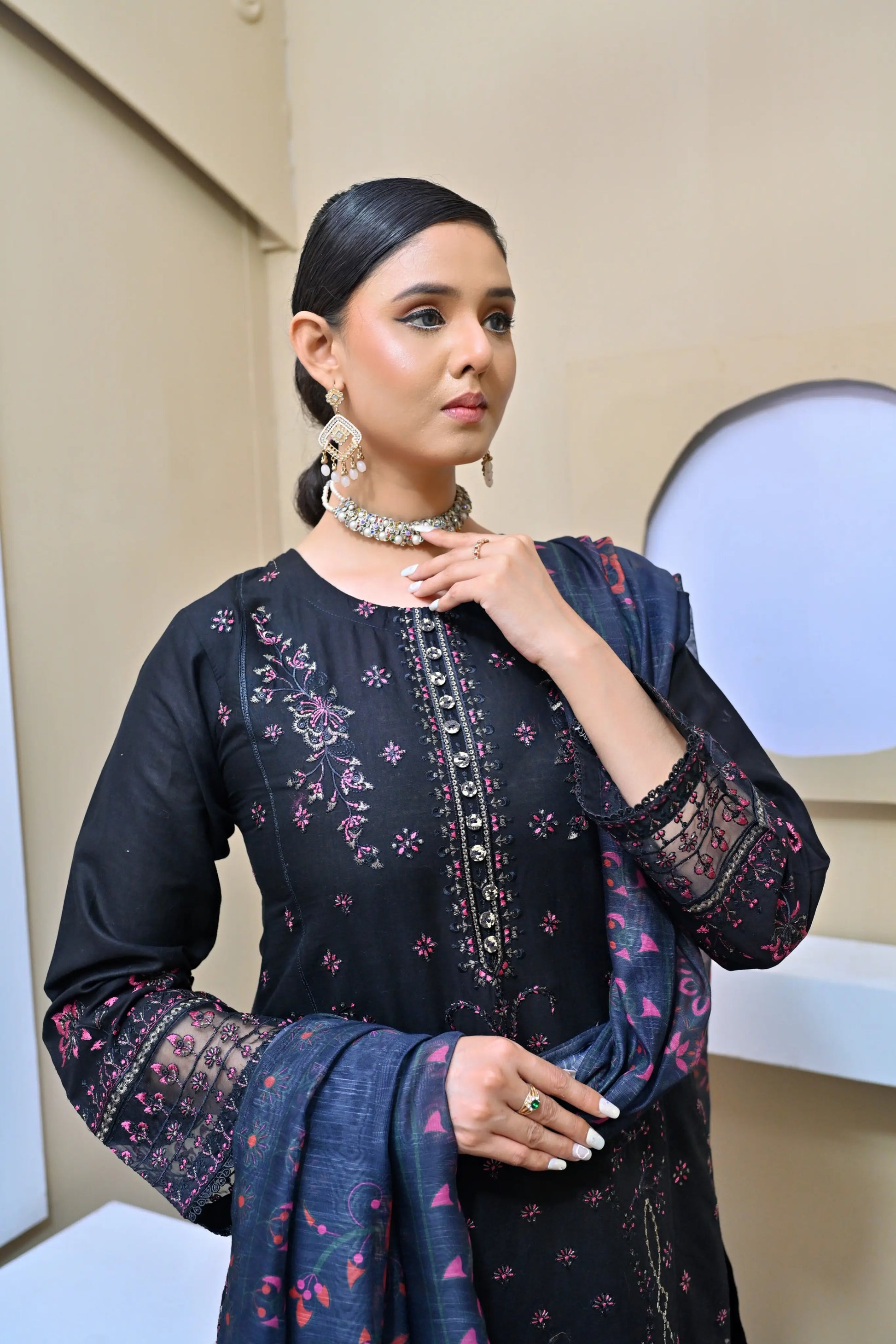 Model in a black three-piece embroidered suit with pink floral details, lace-trimmed sleeves, and a printed dupatta, posing in a minimalist studio setting.