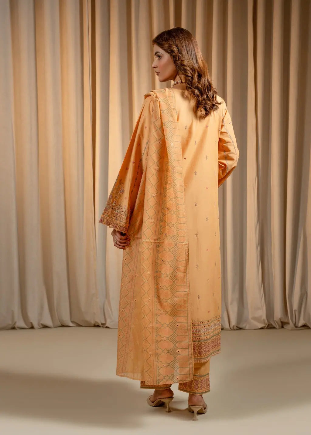 Woman wearing a long, light brown dress with a patterned design against a beige curtain background.