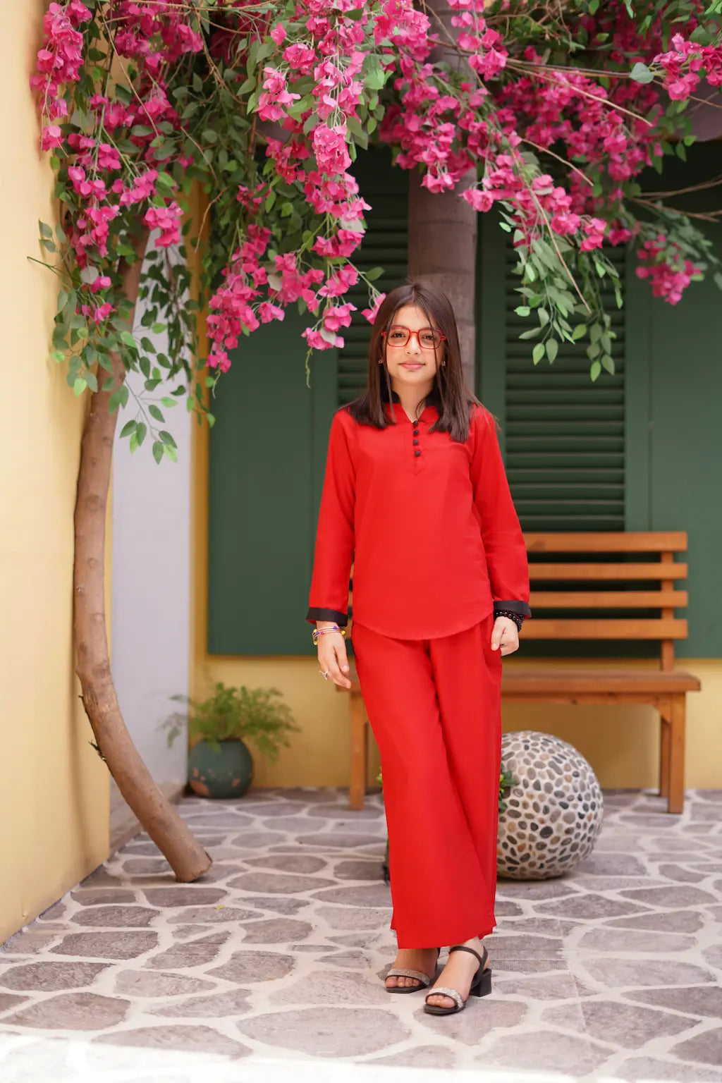 A young girl in a red outfit standing in front of a building with pink flowers and a wooden bench.