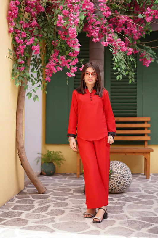 A young girl in a red outfit standing in front of a building with pink flowers and a wooden bench.