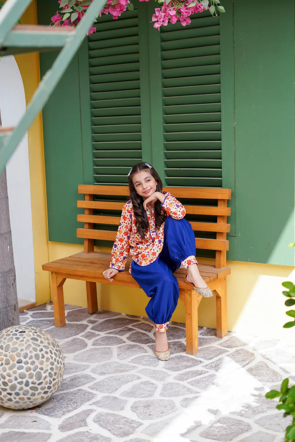 A young girl sitting on a wooden bench in front of a green shuttered building with pink flowers.