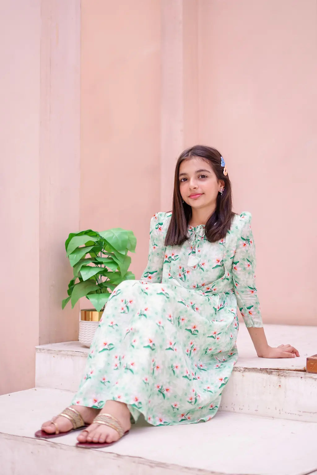 Young girl in a floral dress sitting on steps with a pink wall and plant in the background