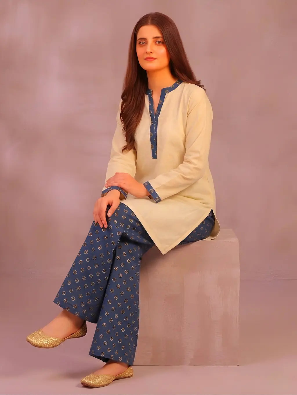 Woman sitting on a box wearing a cream kurta with teel accents and patterned pants against a plain background.