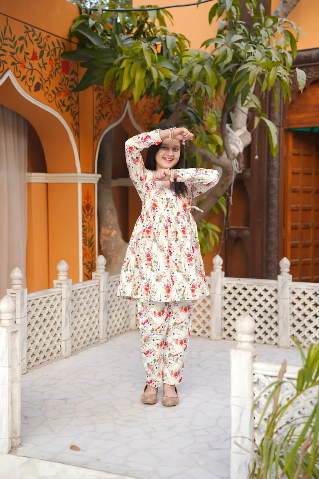 Young girl in a floral outfit posing in a courtyard with orange walls and white lattice.
