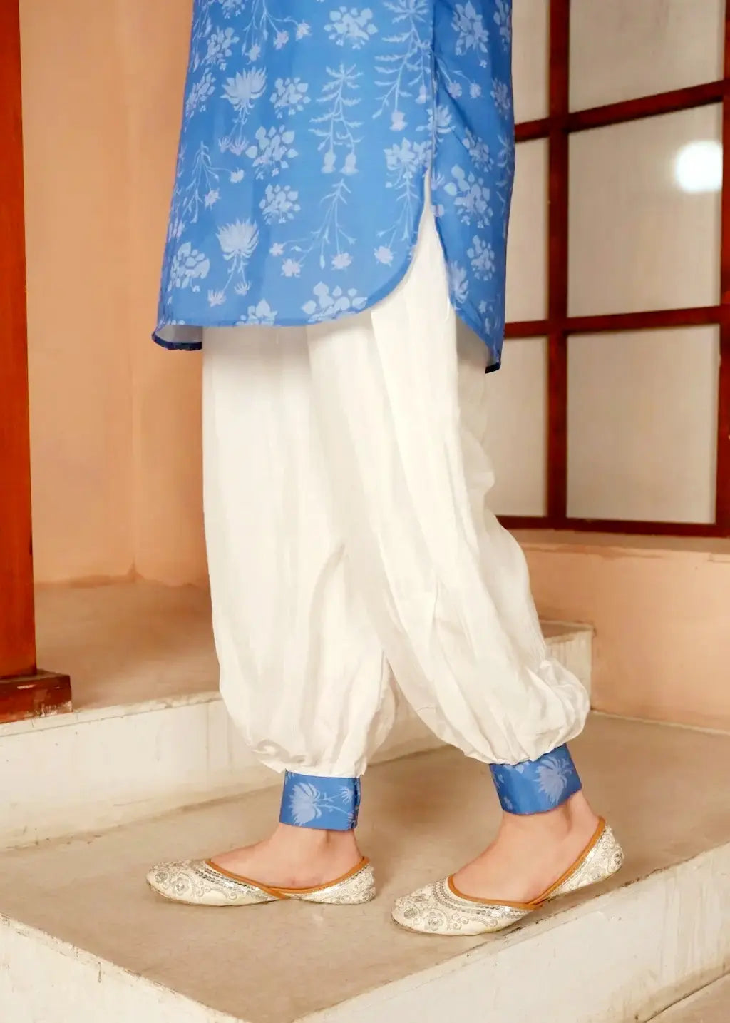 Young girl wearing a blue floral top and white pants with blue cuffs, standing indoors.