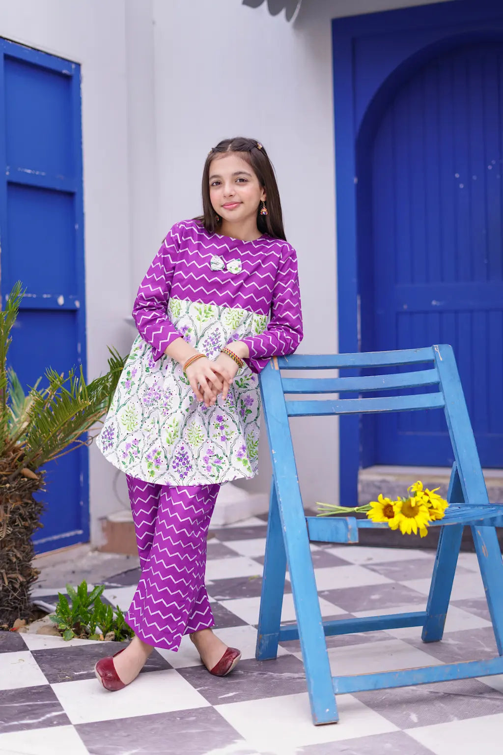 Young girl in a purple outfit standing in front of a blue door with a checkered floor.