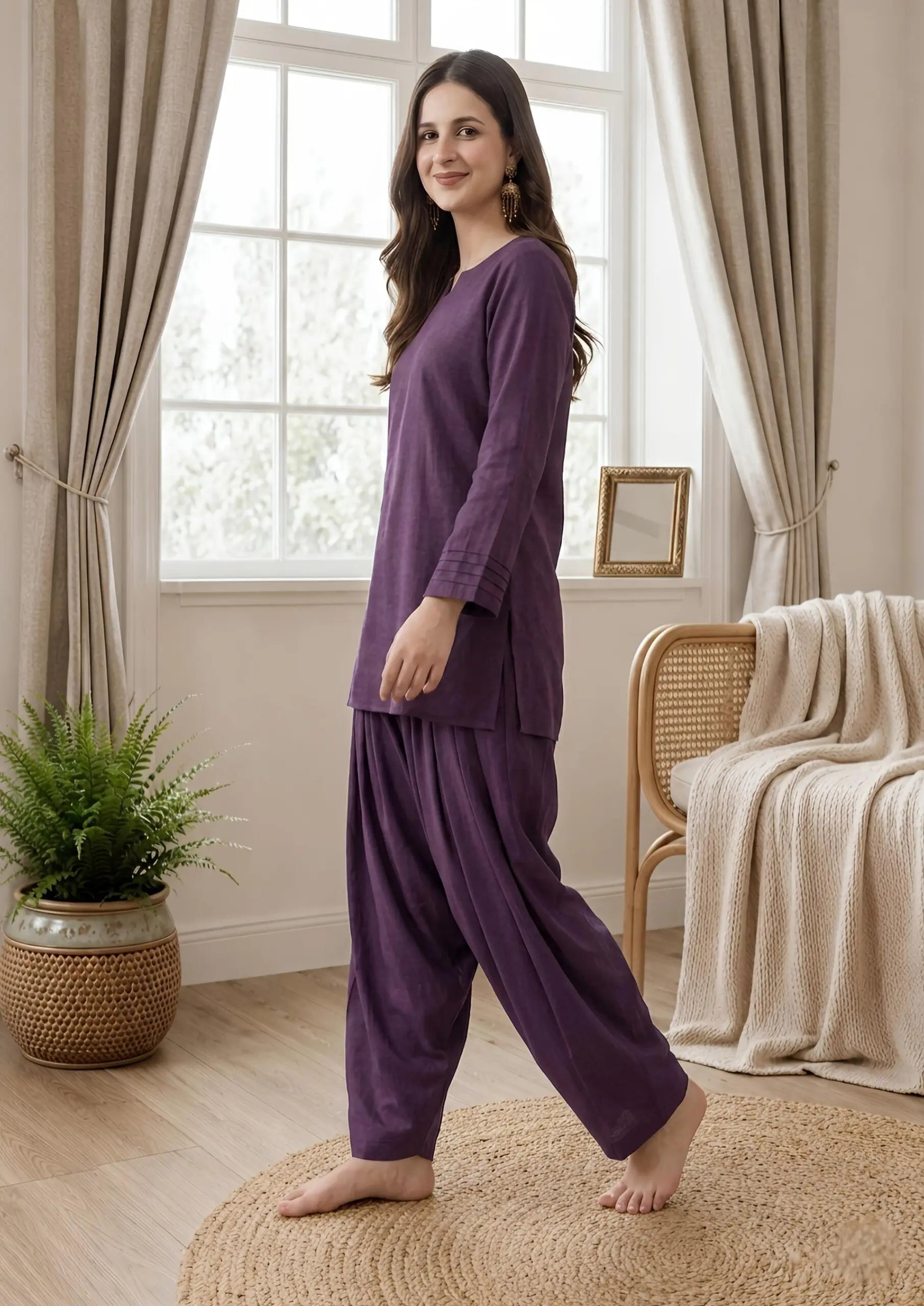 Woman in a purple farshi shalwar set standing in a cozy room with curtains and a plant.