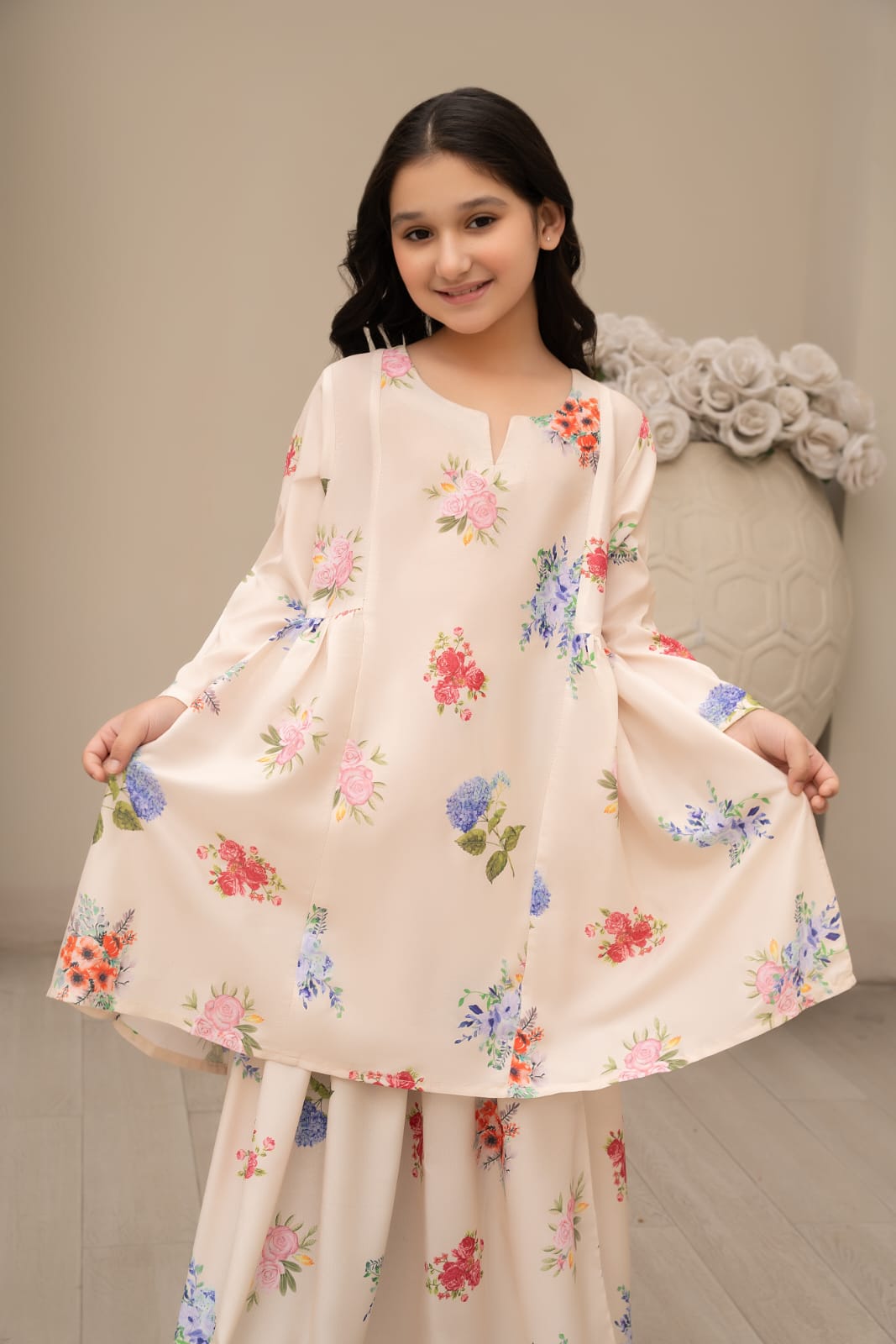 A young girl wearing a traditional cream-colored silk lehnga with floral prints, consisting of a long-sleeve top and a flared skirt.