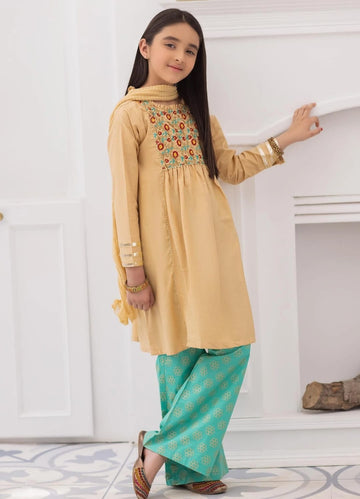 A girl wearing a beige dress with floral embroidery and blue pants standing indoors.